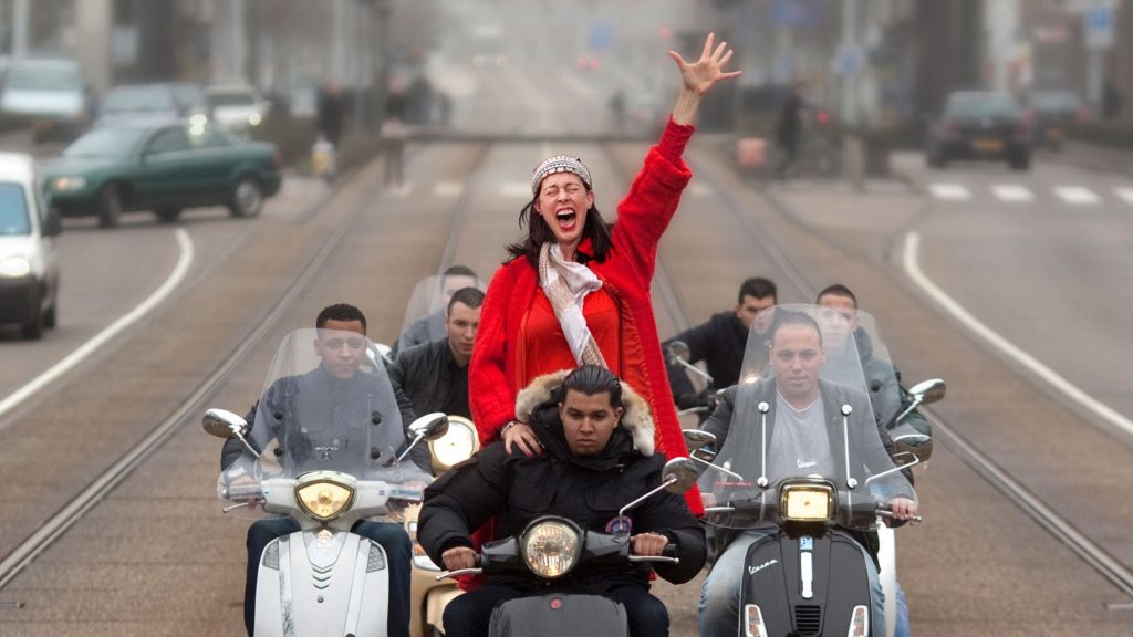 Adelheid Roosen achterop een scooter met rode jas